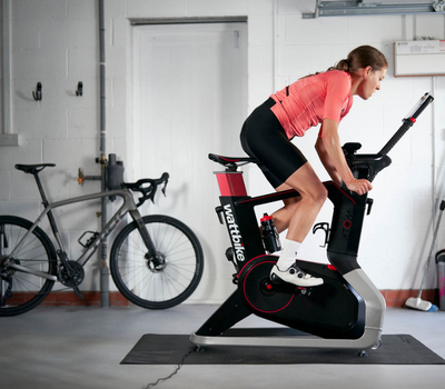a woman in a pink jersey cycles on a Wattbike in her garage with her road bike in the background